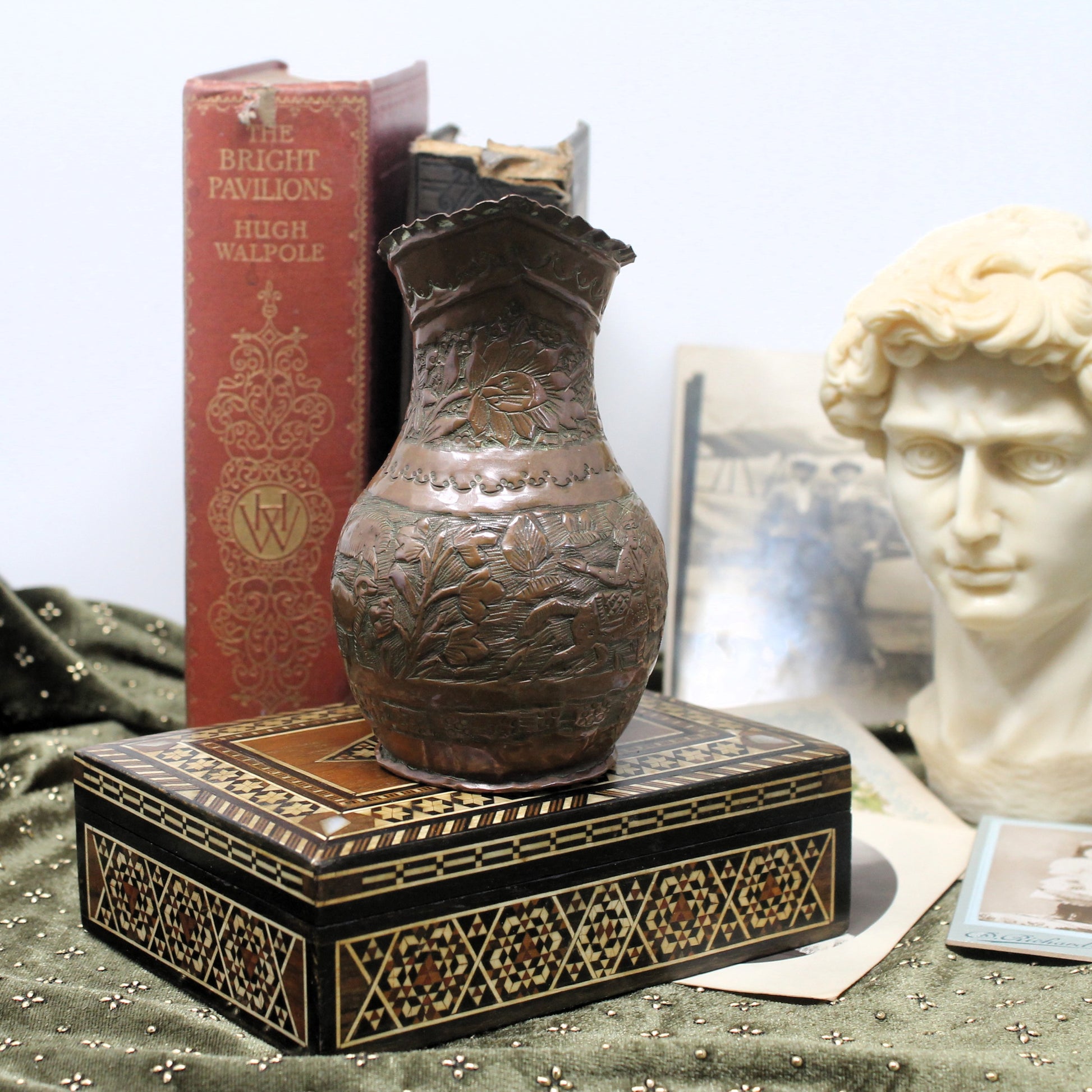 An antique Egyptian carved copper pitcher vase displayed on a black and gold patterned decorative box, with vintage books and a classical bust in the background.