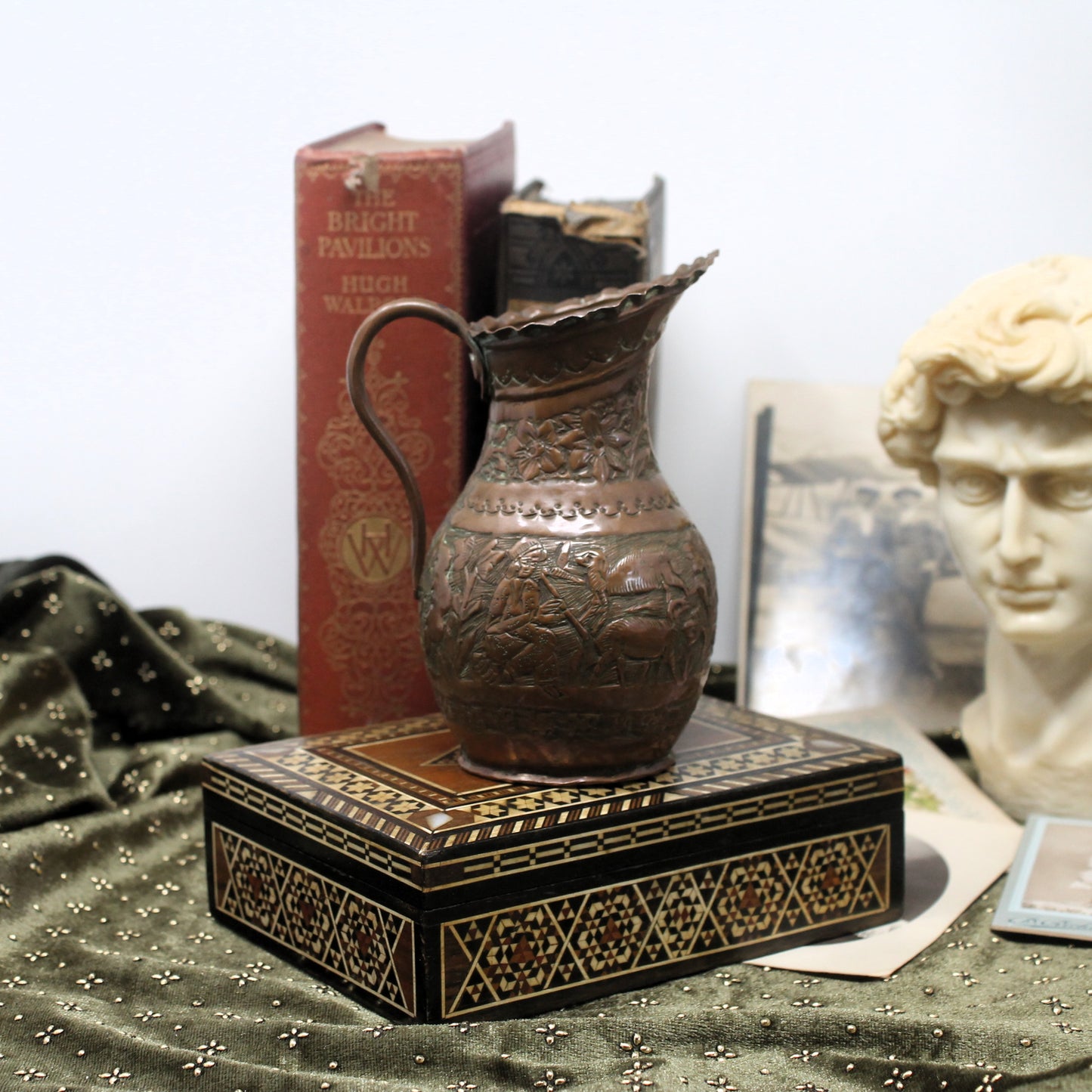 An ornate hammered copper pitcher with a curved handle, displayed on top of a black and gold patterned box. The scene is styled with vintage books, a draped green fabric, and a classical bust in the background.
