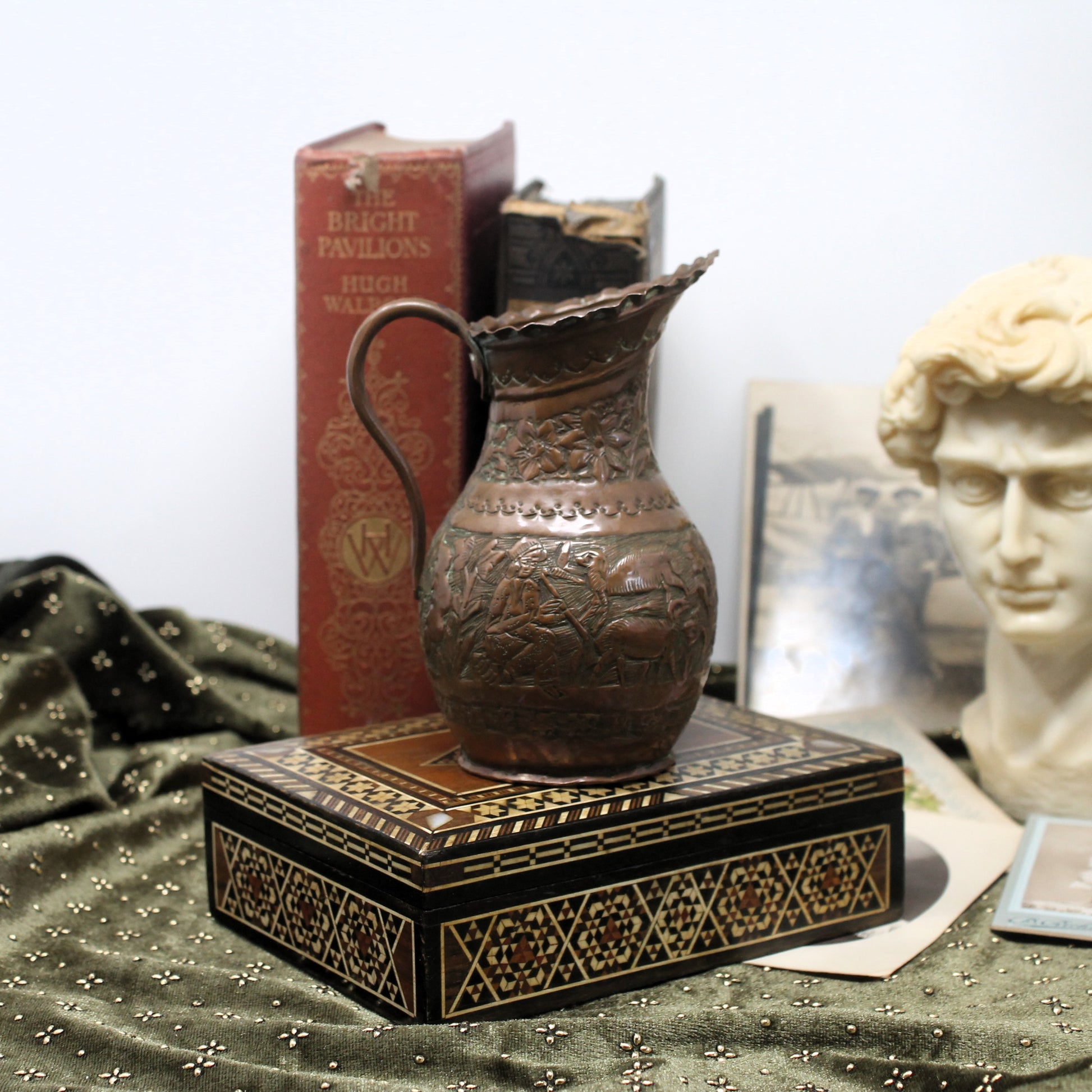 An ornate hammered copper pitcher with a curved handle, displayed on top of a black and gold patterned box. The scene is styled with vintage books, a draped green fabric, and a classical bust in the background.