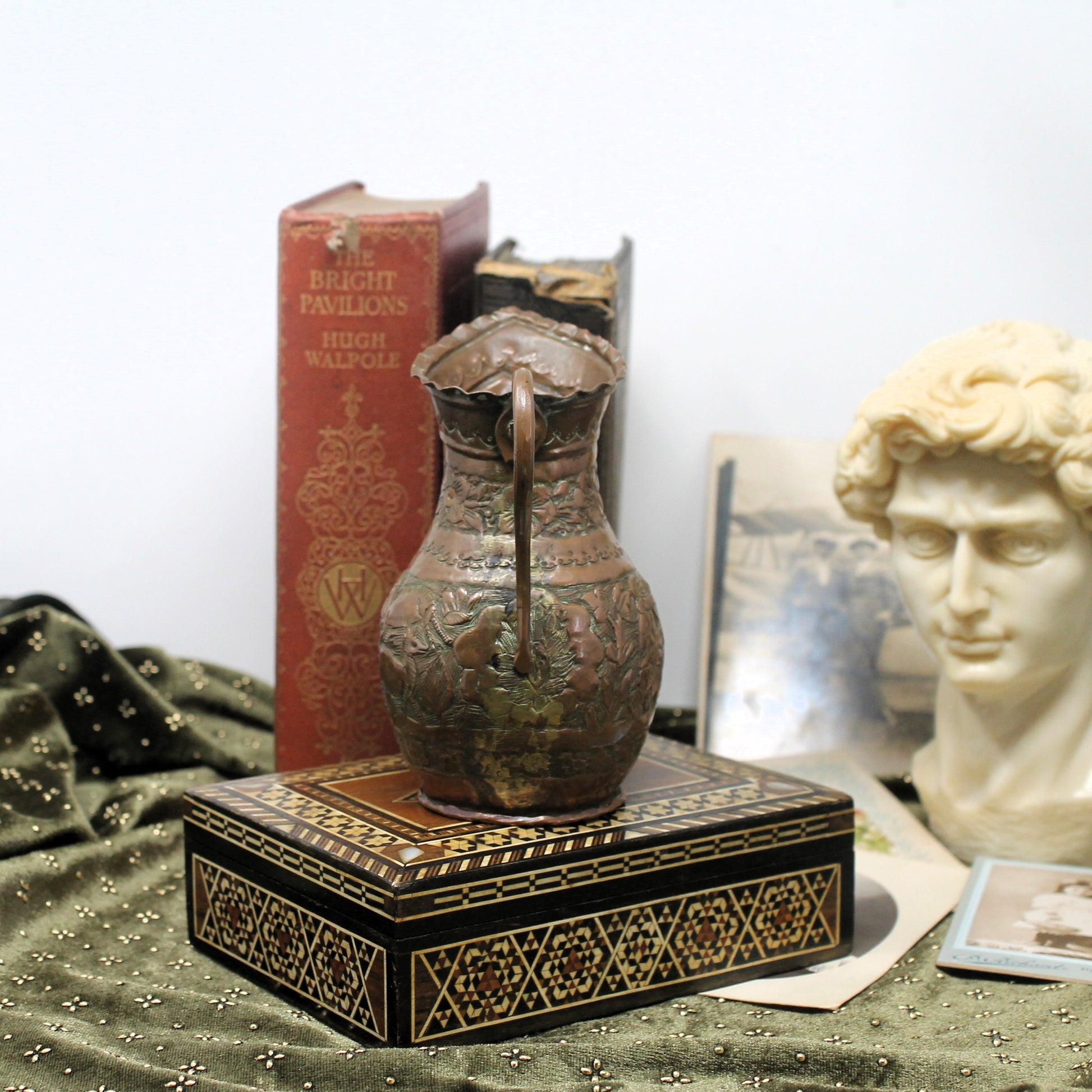 An ornate Levantine antique-style copper pitcher with a handle and decorative relief, displayed on a black patterned box. The scene includes a red book, a classical bust, and framed pictures against a plain light background.