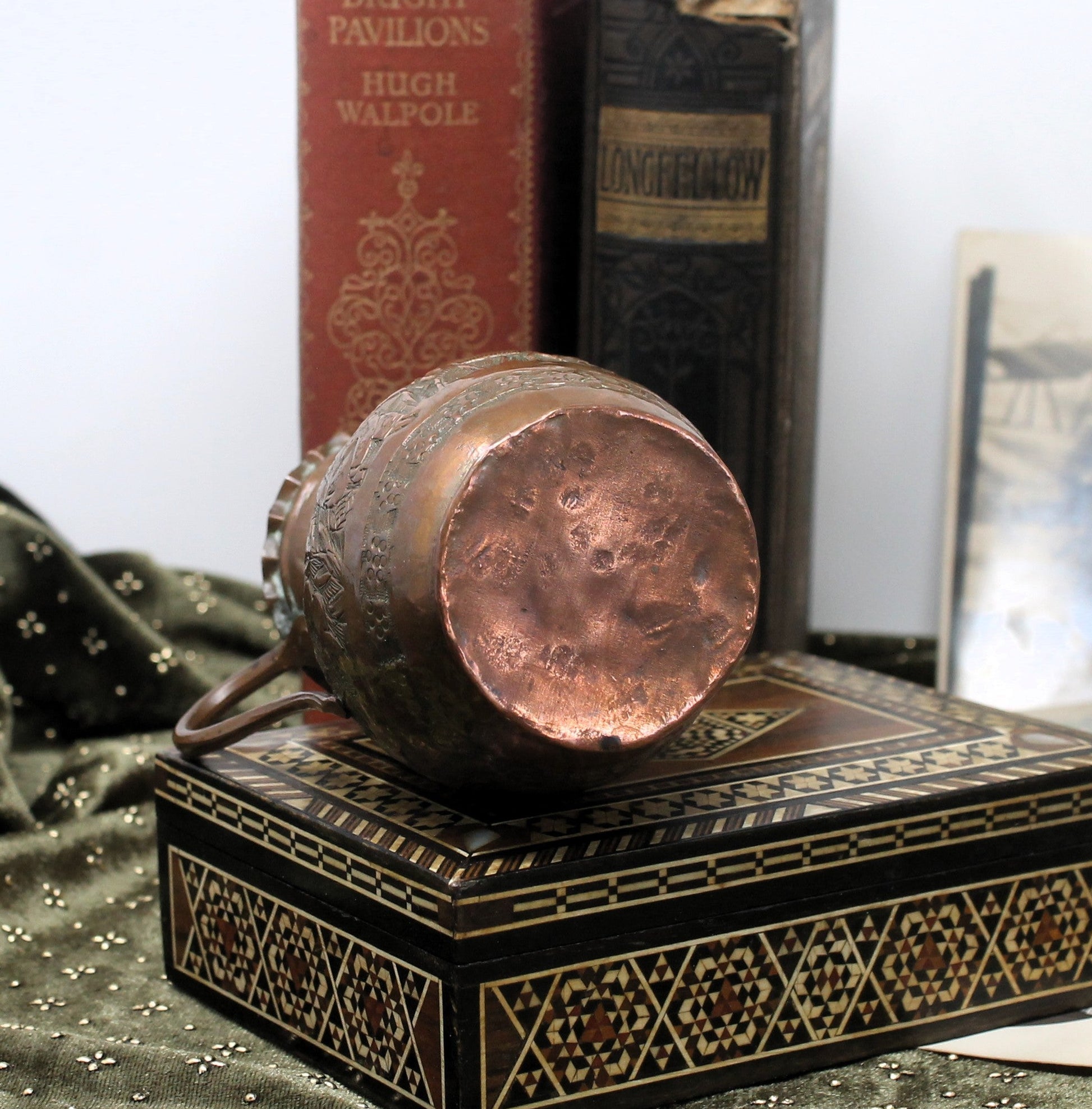 A copper-colored decorative jug with embossed floral patterns, shown lying on top of an ornate black and gold box. Vintage books stand behind it on a white surface, with a dark green patterned fabric draped in the foreground.