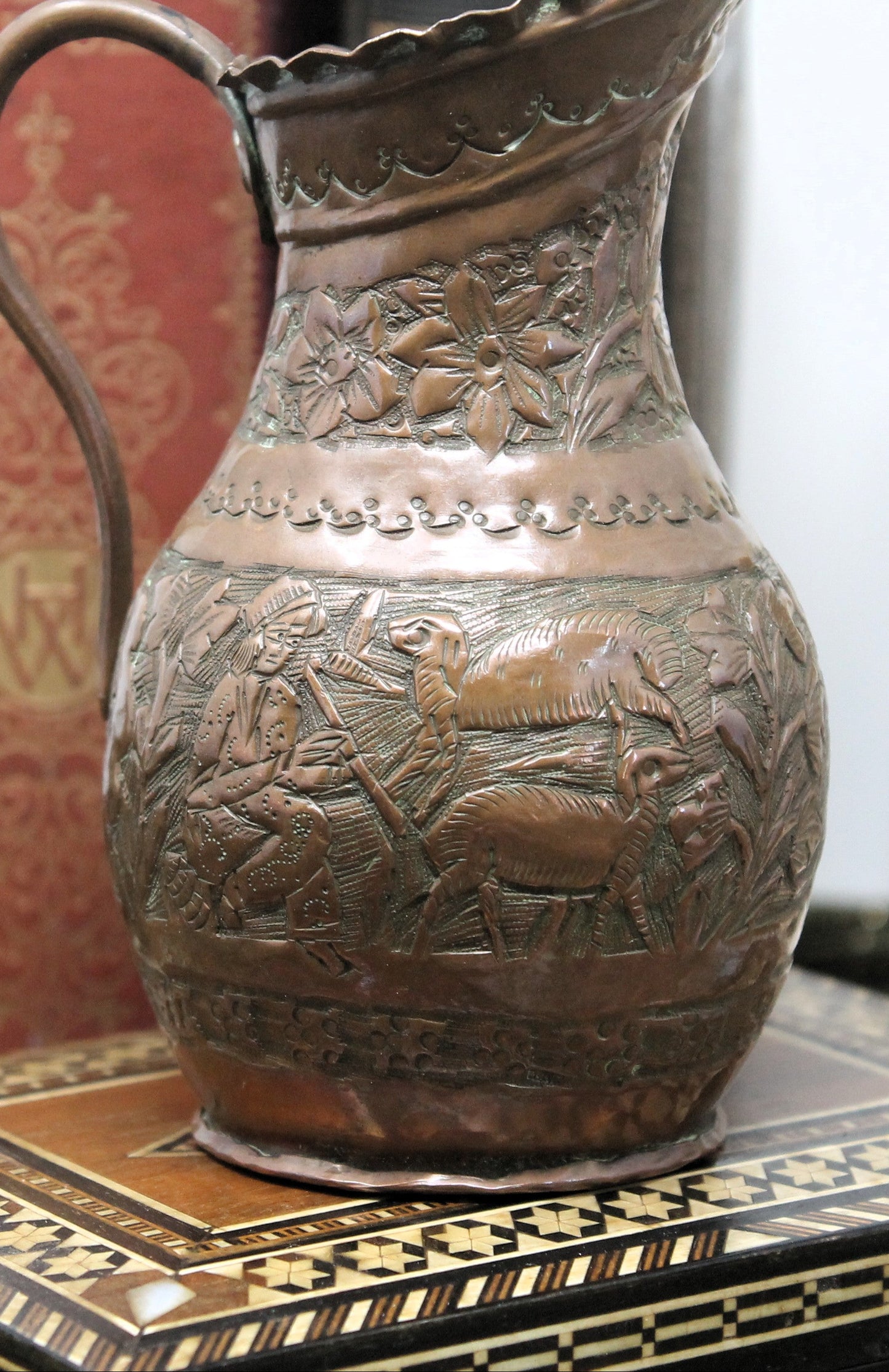 An antique Oriental copper metal pitcher with embossed floral and pastoral relief patterns, displayed on a patterned wooden surface.