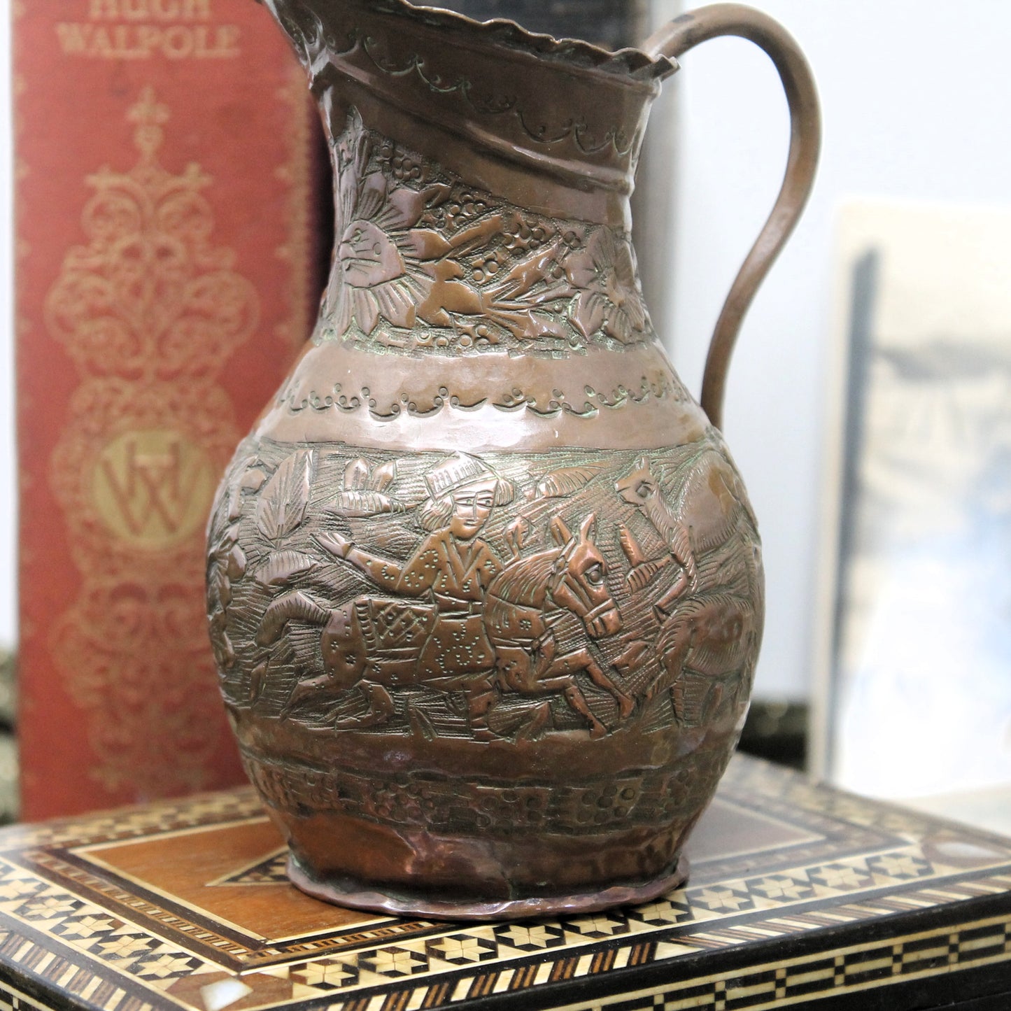 An antique copper hand-hammered pitcher with embossed pastoral scenes, displayed on a patterned wooden surface with a red book in the background.