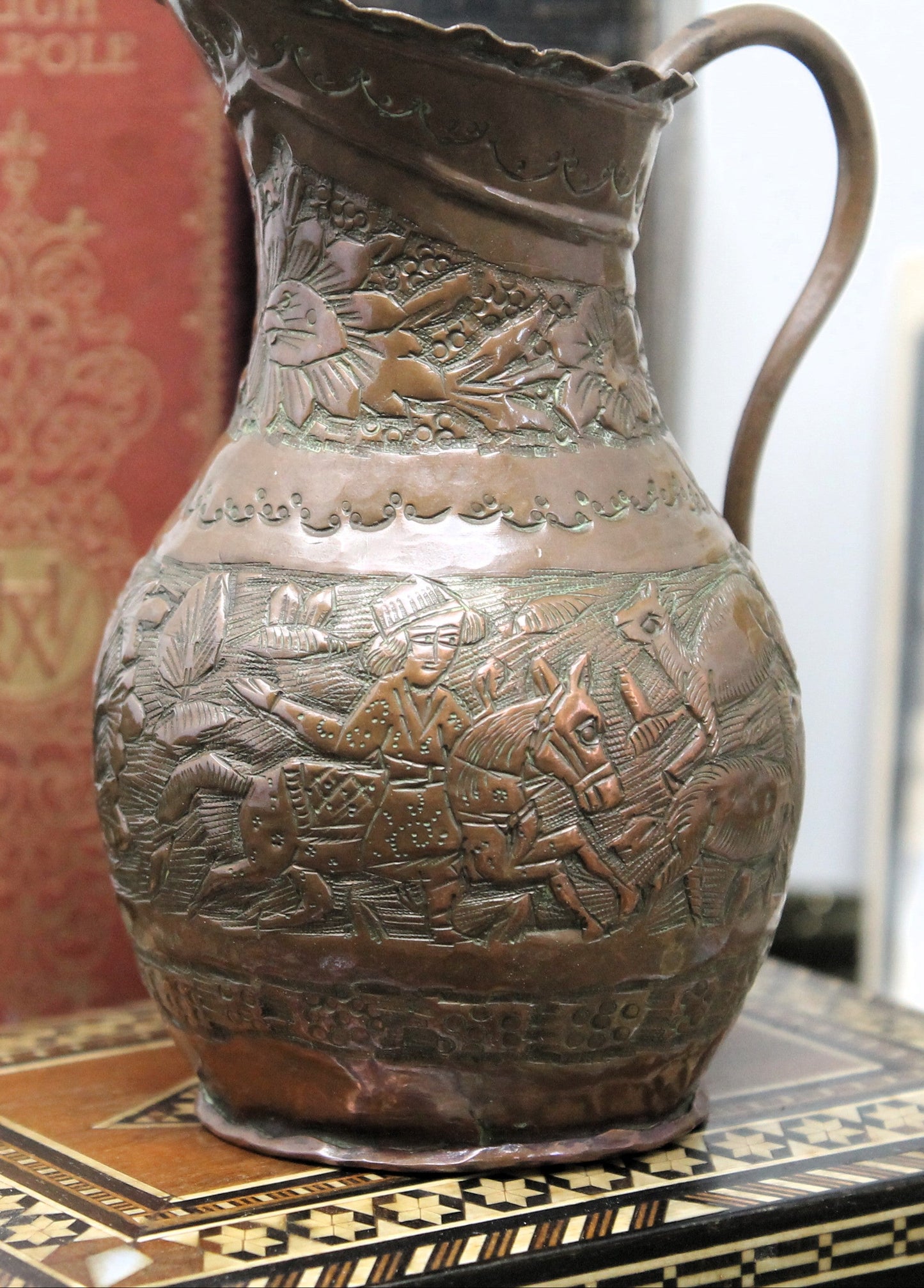 An antique coppe Jerusalem ewer with embossed sheperds scenes displayed on a patterned wooden surface with a red book in the background.
