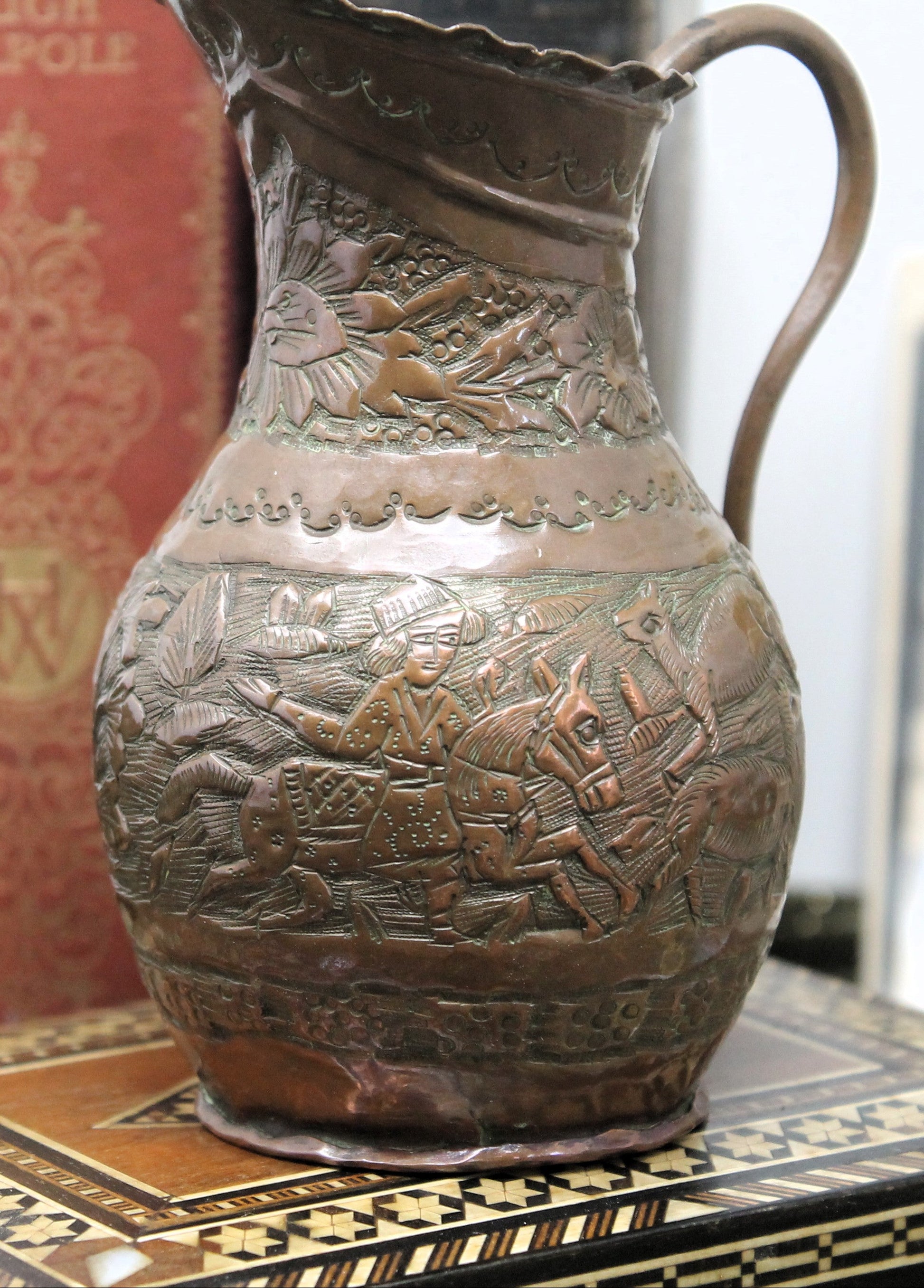 An antique coppe Jerusalem ewer with embossed sheperds scenes displayed on a patterned wooden surface with a red book in the background.