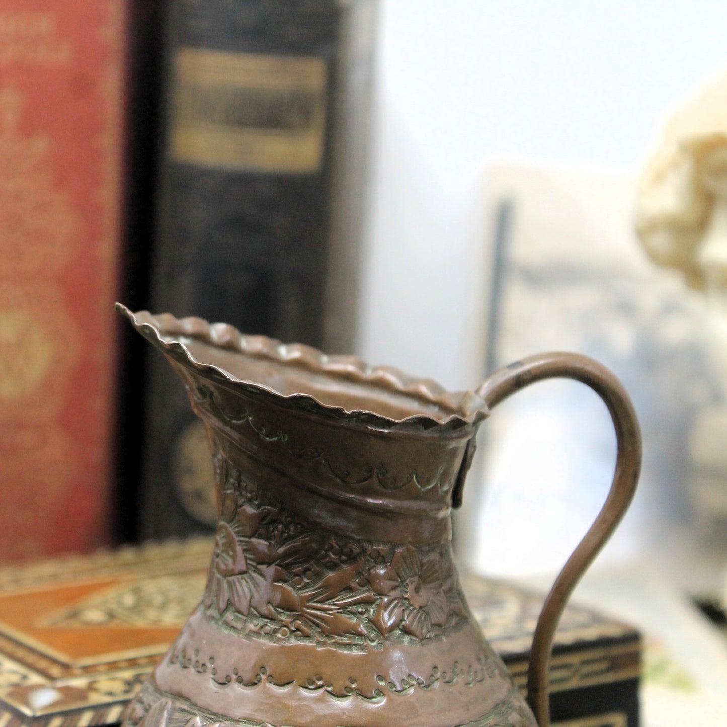 An antique-style moorish copper or bronze pitcher with embossed floral patterns and a flared rim, shown in a close-up shot with a blurred background of books and a bust. 
