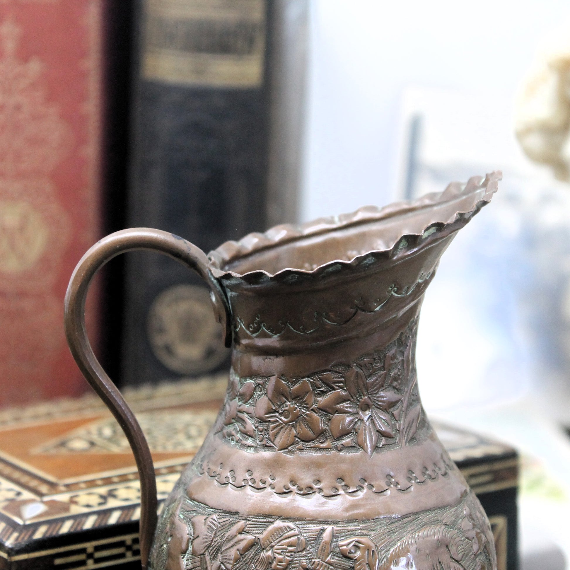 An ornate Middle-Eatern copper pitcher with embossed floral and animal motifs, shown in a close-up setting with blurred books in the background.