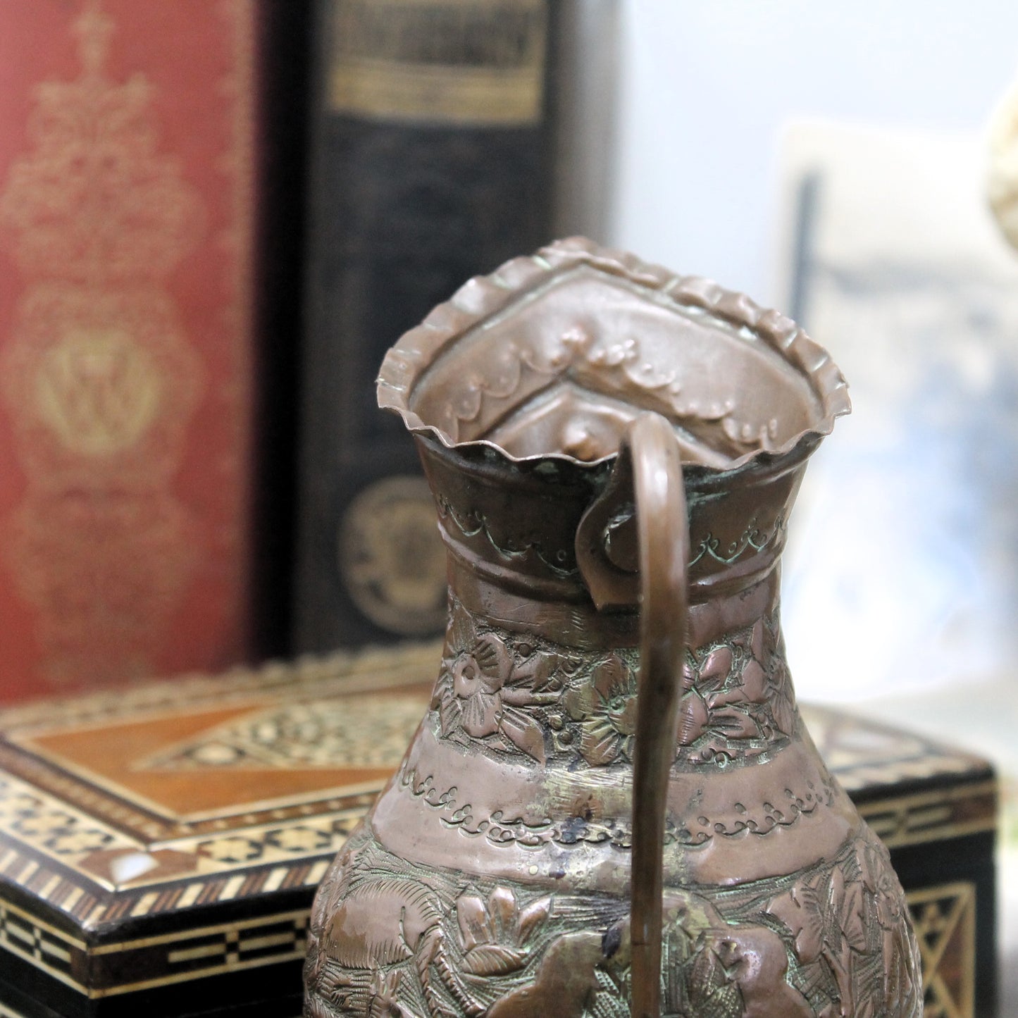 An ornate embossed copper pitcher with a ruffled rim and loop handle, shown in close-up with a blurred background of books and decorative boxes.