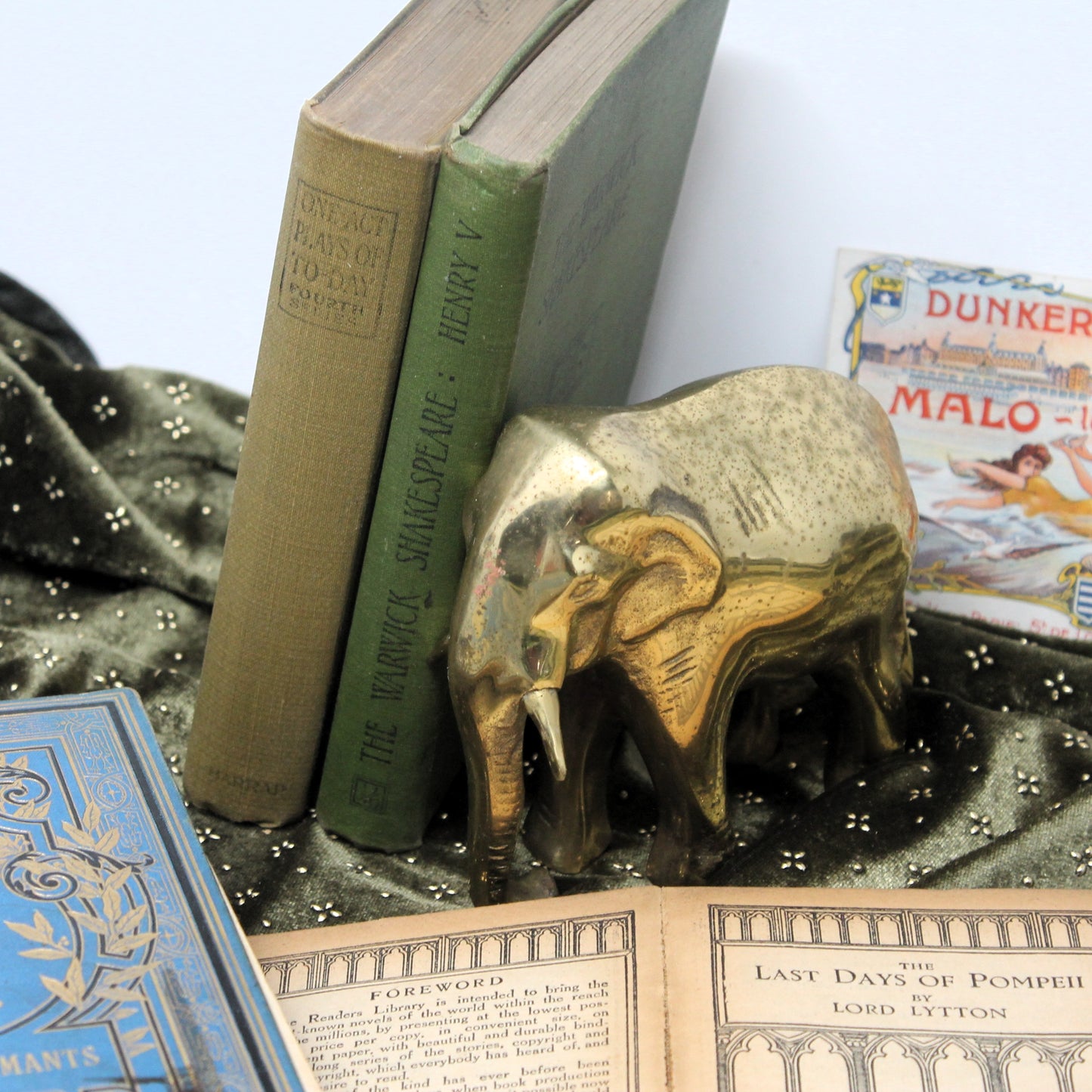 A decorative brass elephant figure bookend holding two vintage books on a dark patterned fabric surface, with additional books partially visible around it.