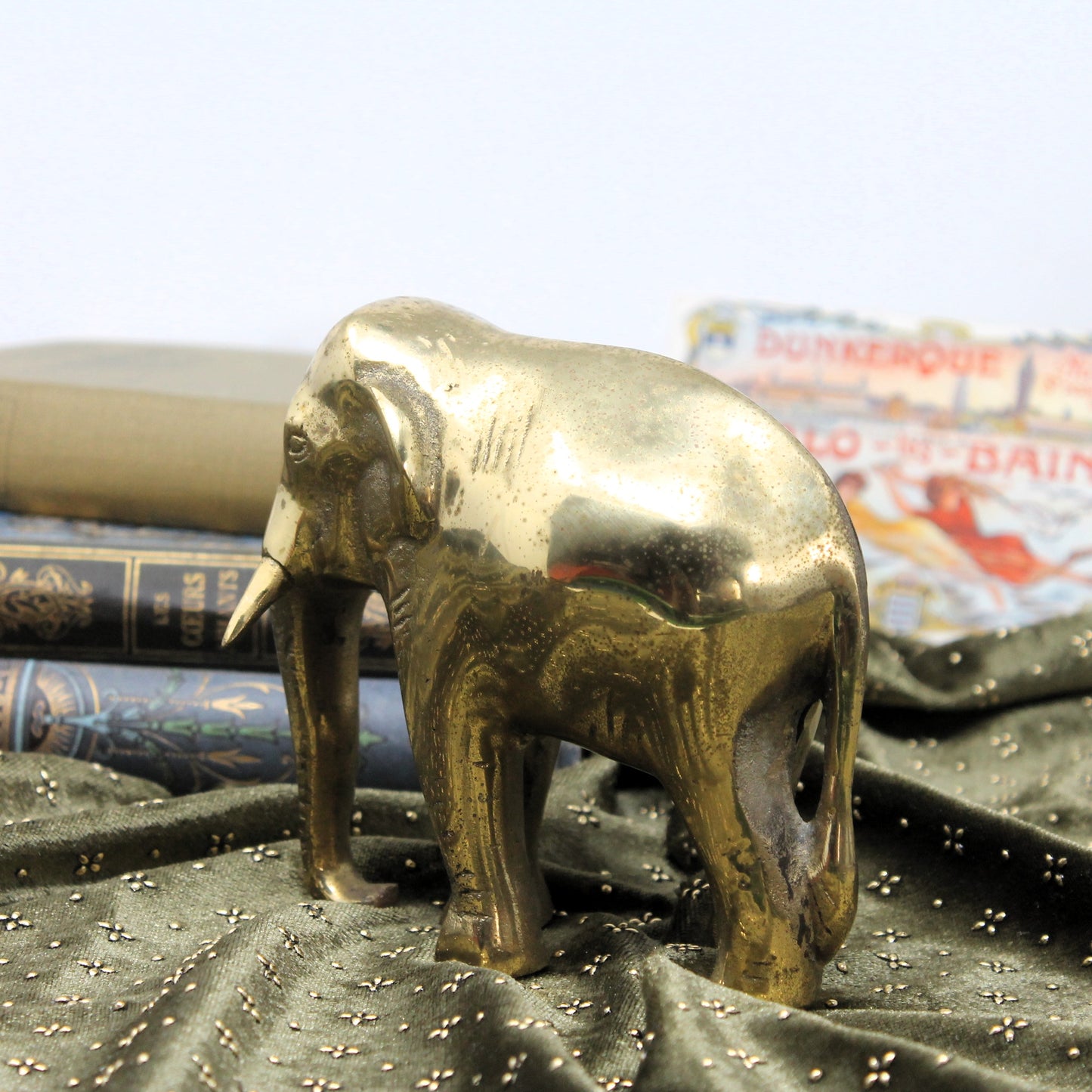 Mid-century Indian or Nepalese elephant figurine with a polished brass finish, standing on dark draped fabric with vintage books and a blurred poster in the background.