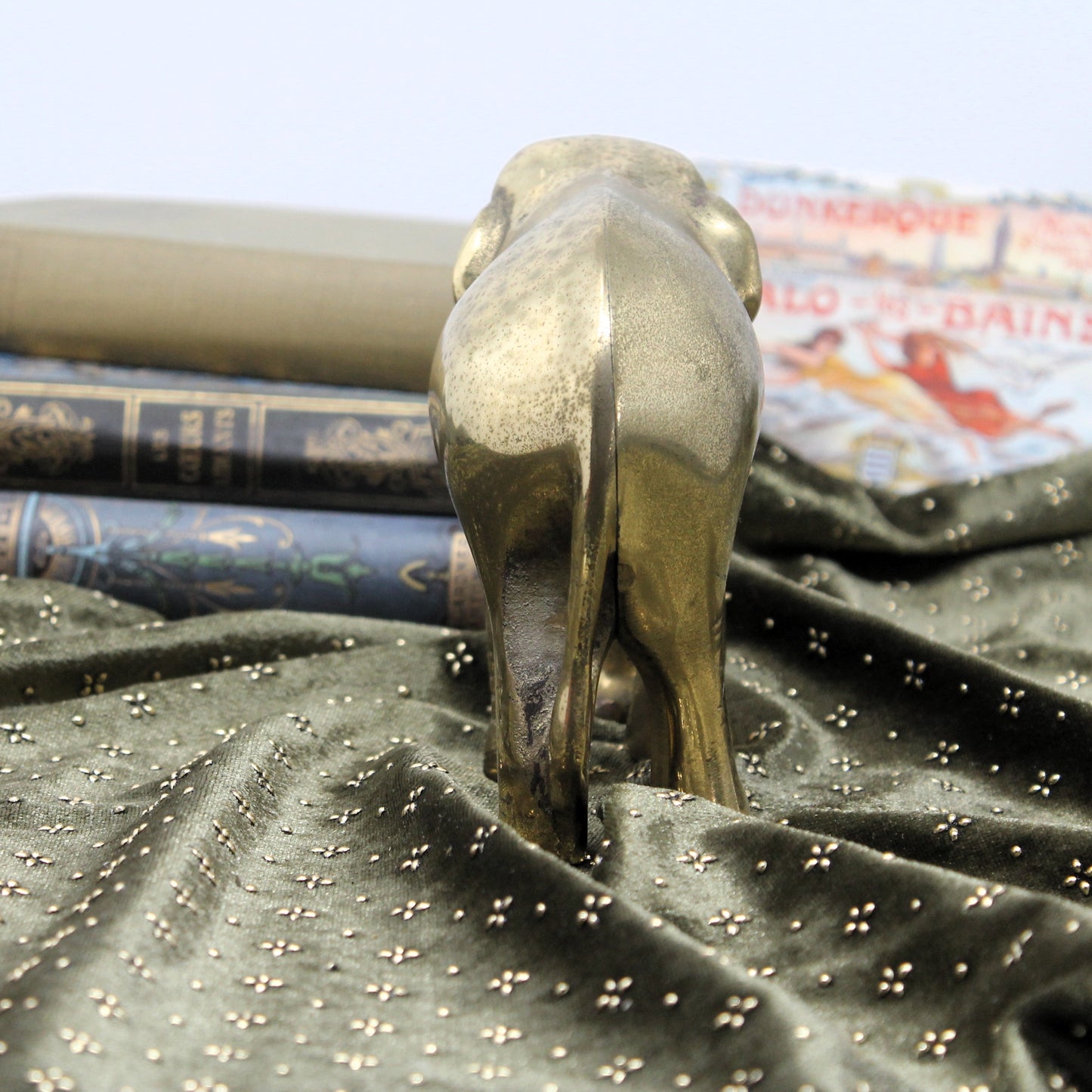 A polished brass elephant figurine standing on its hind legs, positioned on a dark green velvet fabric with small gold star details. In the background, there are stacked antique-style books.