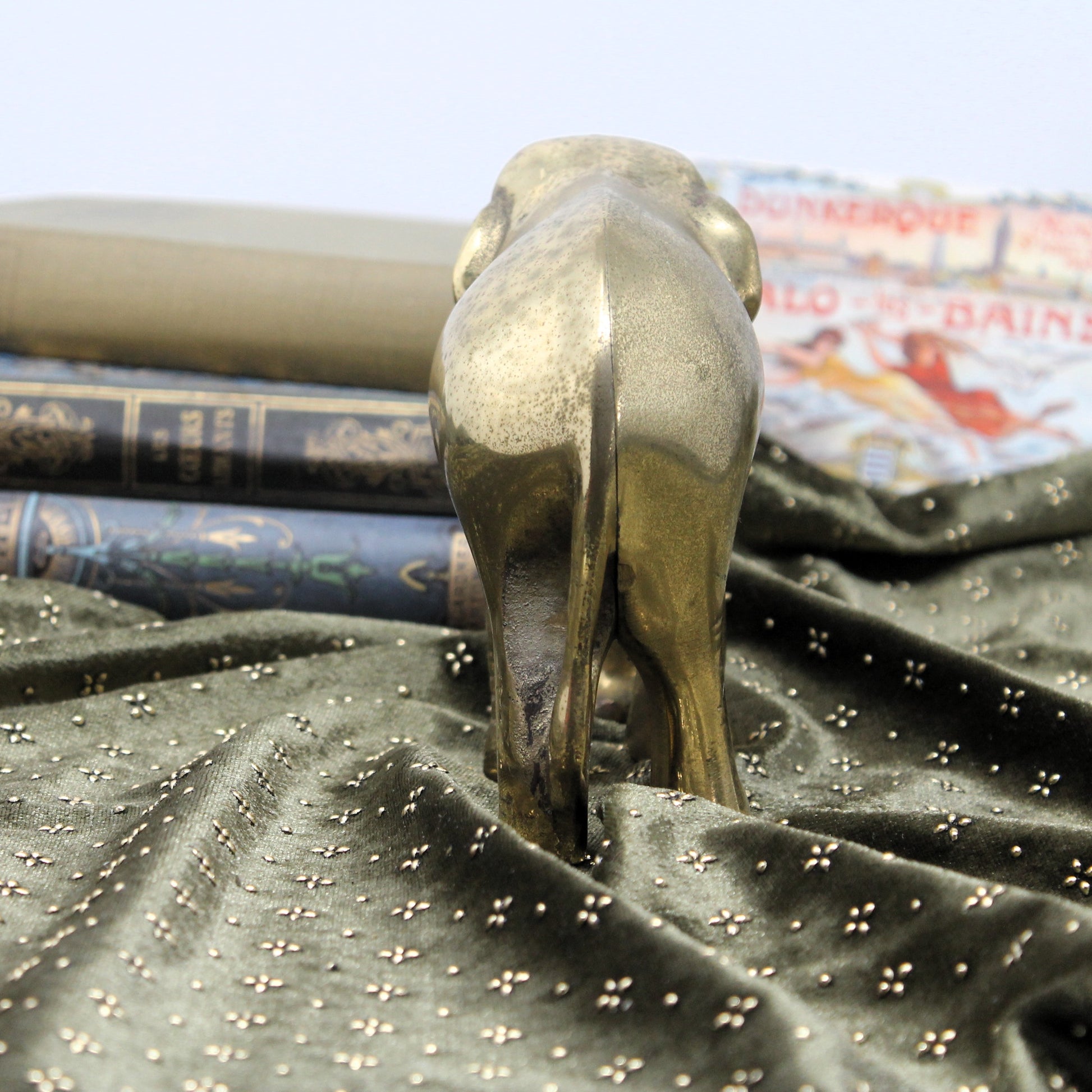 A polished brass elephant figurine standing on its hind legs, positioned on a dark green velvet fabric with small gold star details. In the background, there are stacked antique-style books.