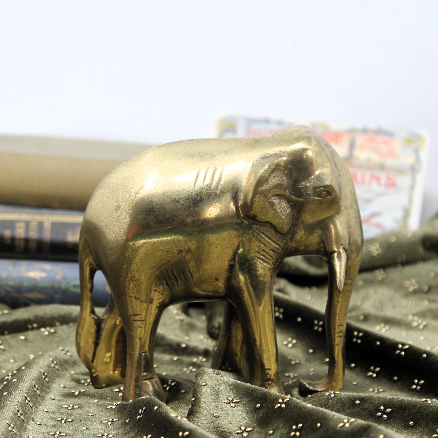 A small metallic elephant figurine in a polished brass or gold finish, standing on a dark draped fabric with small metallic star details. Books are blurred in the background against a pale sky-like backdrop.