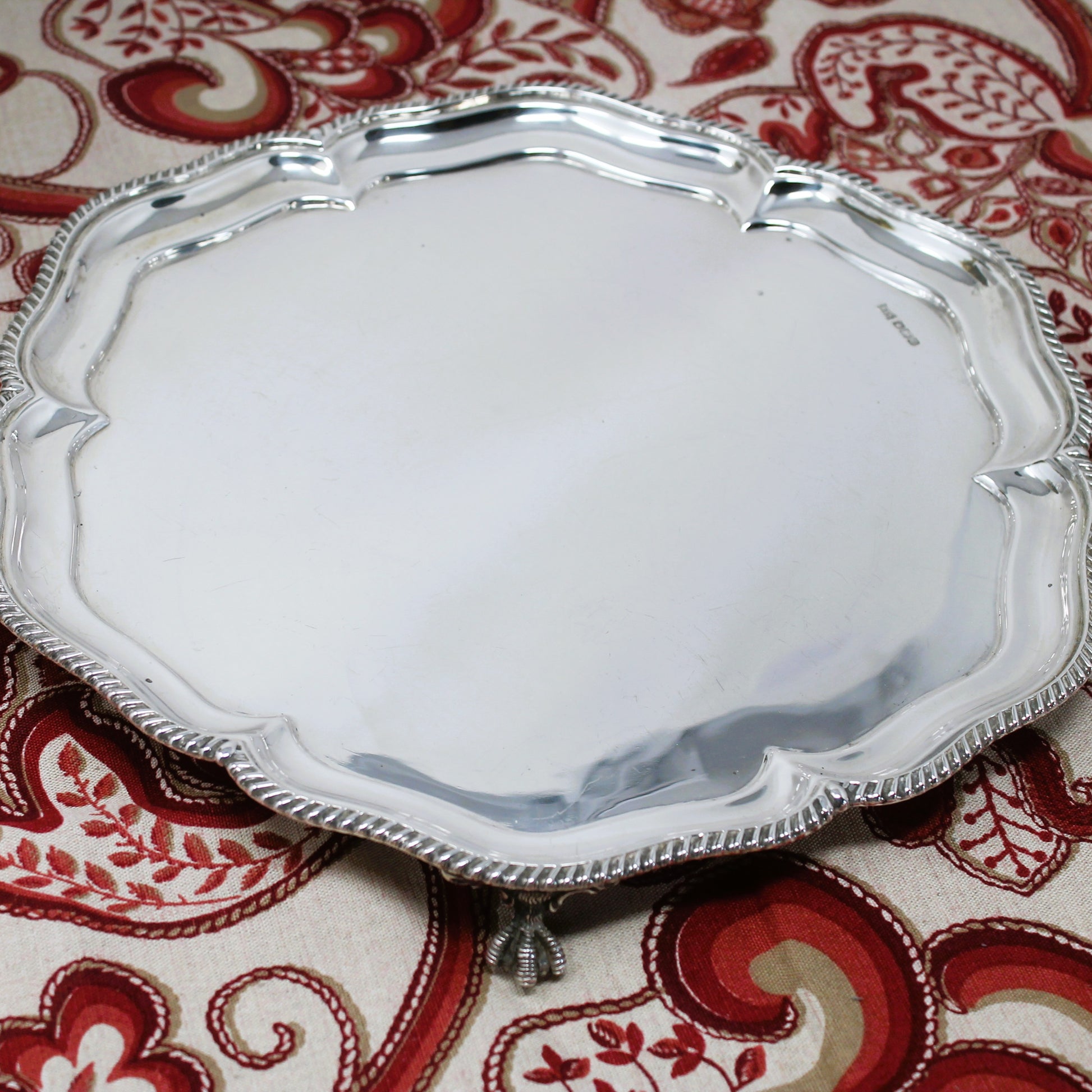 A round silver serving tray with a decorative scalloped edge and small feet, shown on a red and beige paisley patterned fabric background.
