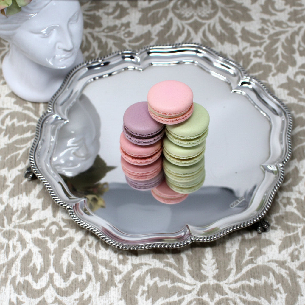 A round sterling silver decorative tray with a scalloped beaded edge, displayed on a beige damask-patterned fabric surface next to a white ceramic bust.