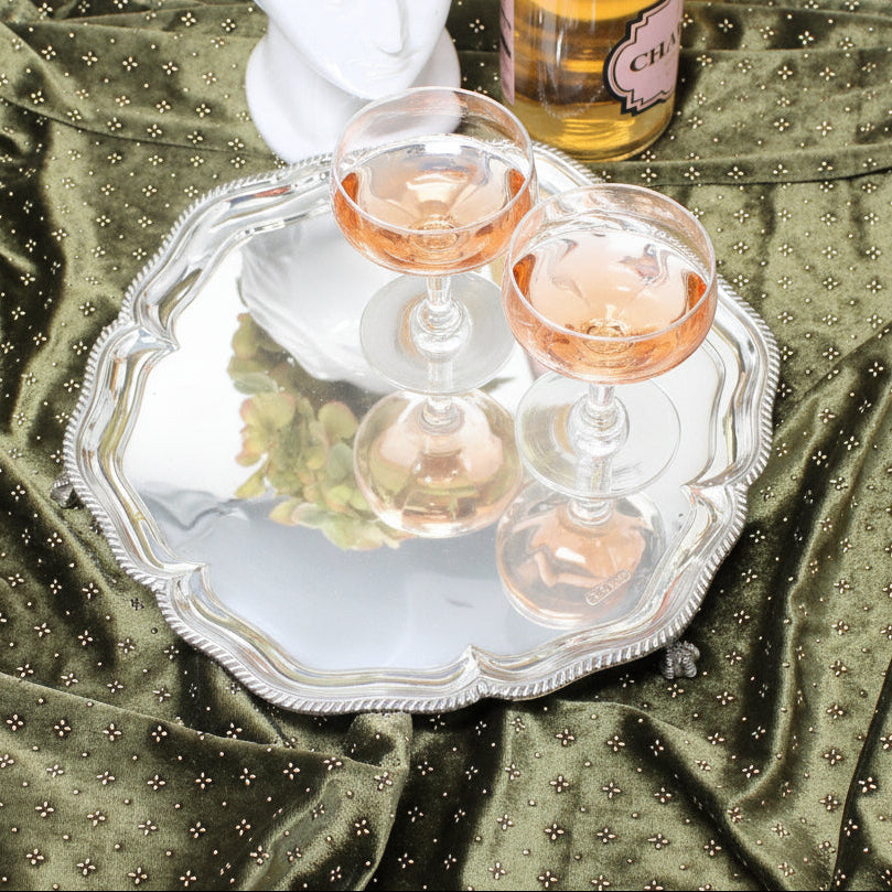 A round silver salver with a scalloped edge, holding two clear stemmed glasses, set on draped olive green satin fabric with small metallic dots.