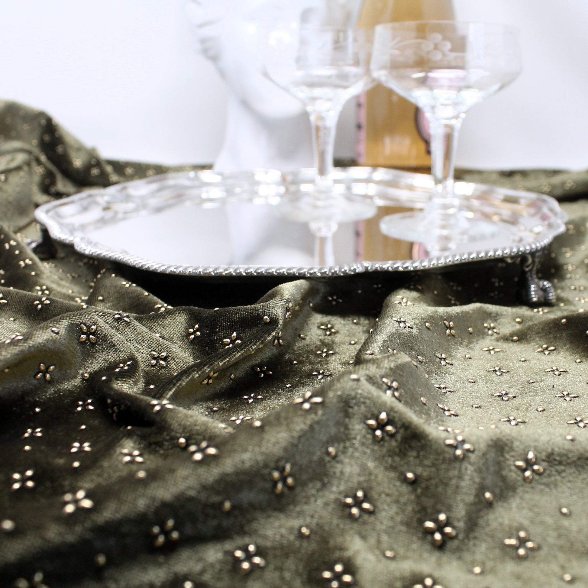An ornate silver tray resting on draped olive green fabric adorned with small metallic floral embellishments, with two champagne glasses and a white decorative bust in the background.