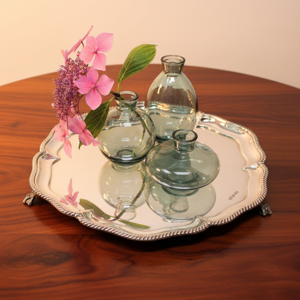 Three small clear glass vases with bulbous rounded vases and narrow necks, arranged on a sterling silver tray by Walker & Hall resting on a mahogany table
