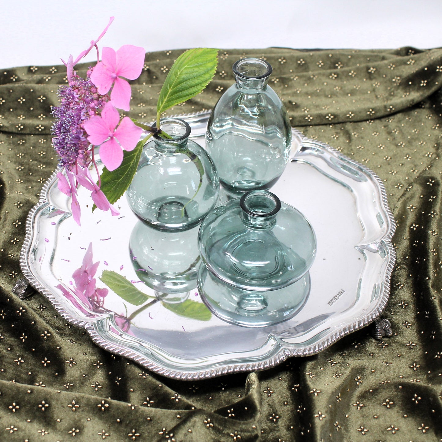Three small clear glass vases with bulbous rounded bodies and narrow necks, arranged on a Sheffield Georgian revival silver tray over an olive green patterned fabric backdrop, with pink flowers placed beside them.