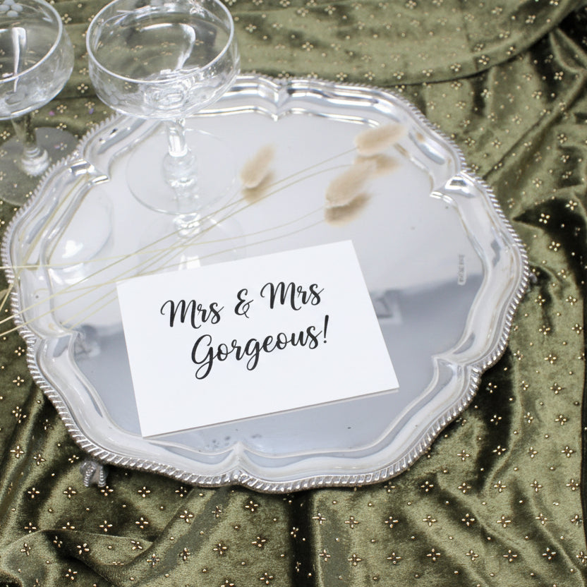 A round silver serving tray with a scalloped edge and beaded rim, displayed on a shiny olive green satin fabric. Two clear stemmed glasses and a few dried stems rest on the tray, which also has a blank white card placed on top.