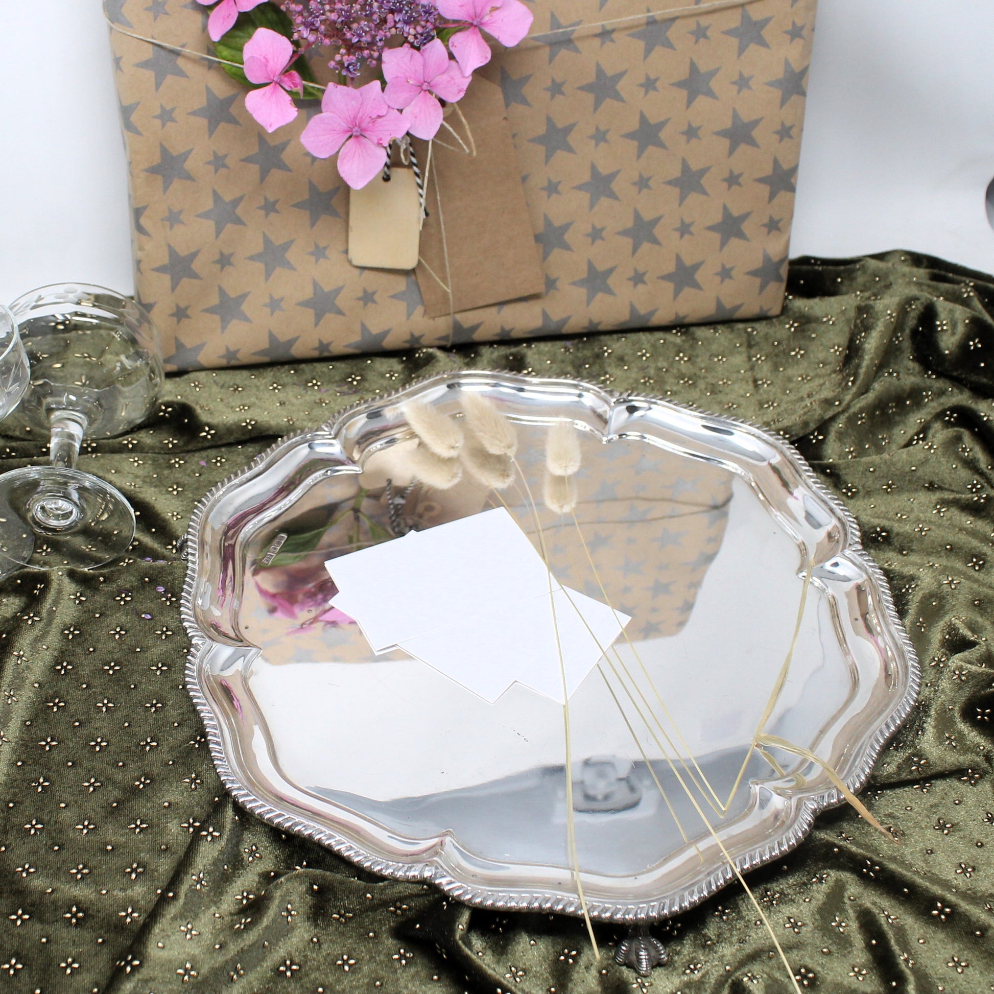 A scalloped silver metal serving tray with a beaded rim, displayed on an olive green dotted fabric. In the background, a kraft paper gift box with a gray star pattern and pink flowers is visible.