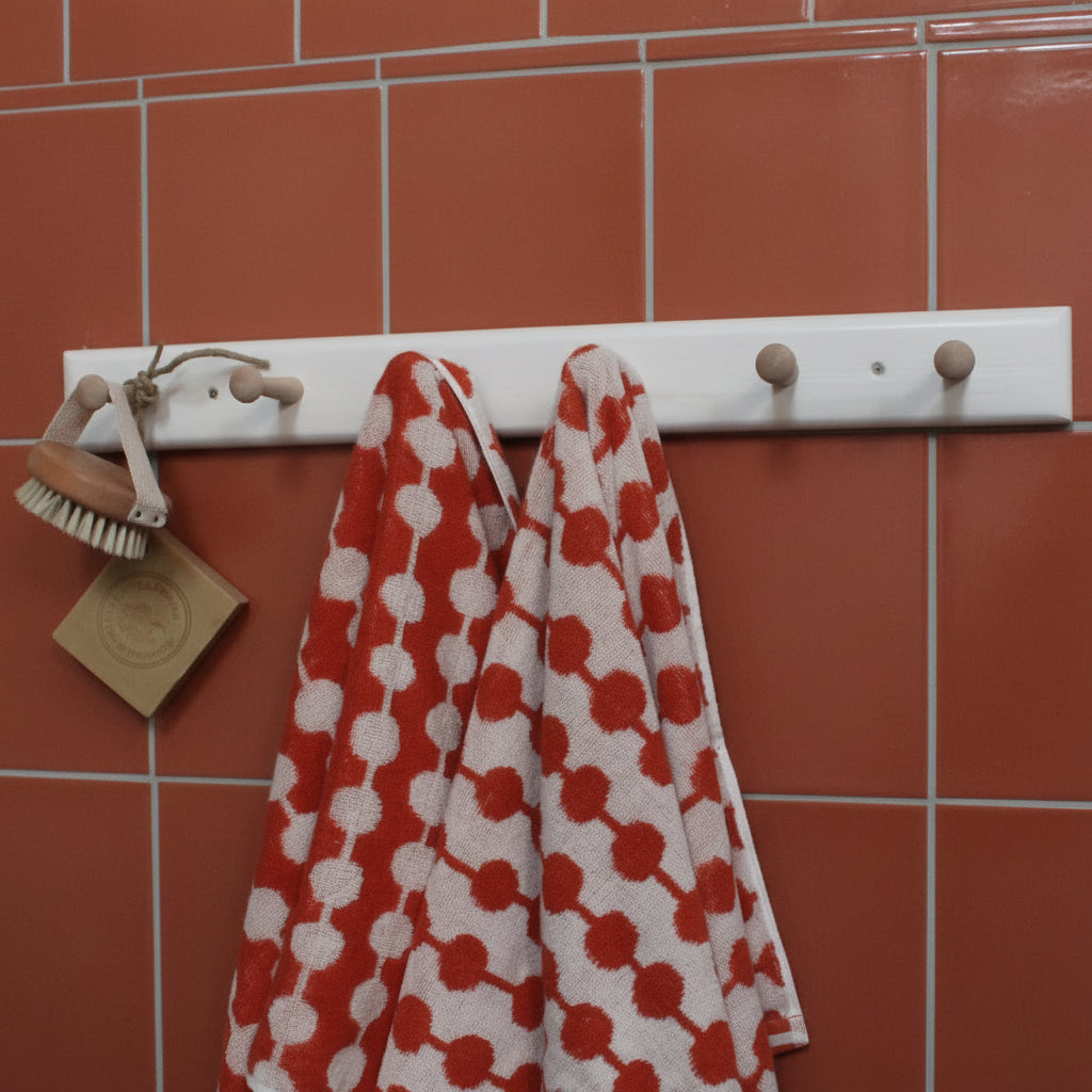 A white wall-mounted hook rack with wooden pegs, holding a red and white patterned towel and a small hanging brush, against a terracotta tiles walll.