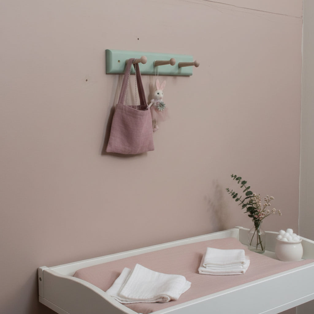 A mint green wall-mounted wooden peg rack with three round pegs, holding a dusty pink fabric bag and a small bunny decoration against an pink wall.