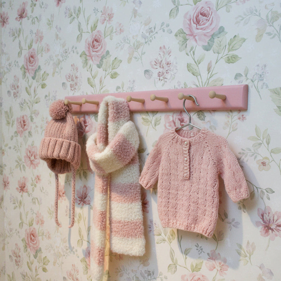 A pink wall-mounted wooden coat rack with eight light natural wood pegs, shown against an off-white wall.