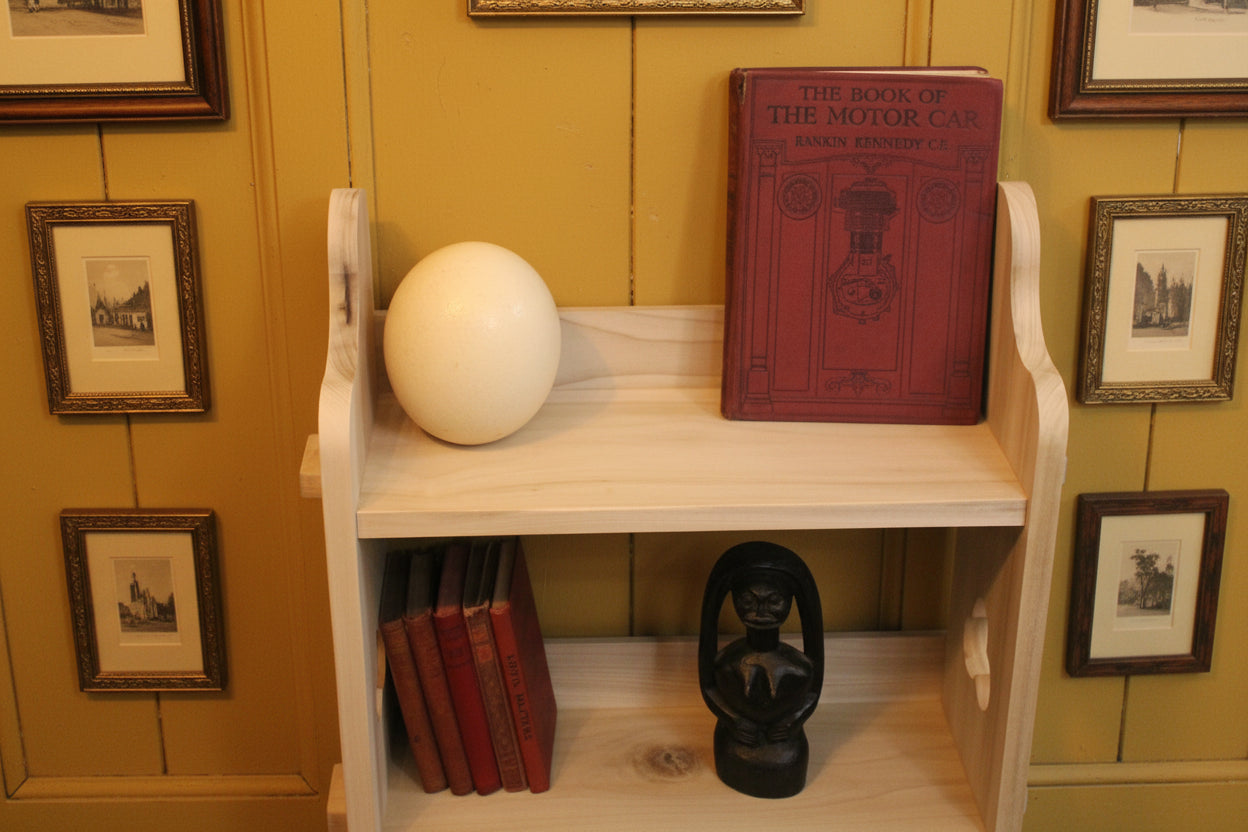 A light natural wood bookshelf styled with a red vintage book, a white decorative sphere, a stack of red books, and a black lantern figurine, set against a yellow paneled wall with framed pictures.