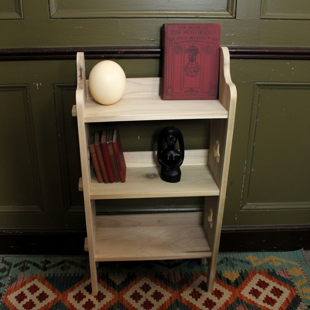 A small light wood three-tier bookshelf styled with a red book, a cream-colored sphere, a stack of red books, and a black lantern figurine, set against a cardboard backdrop.