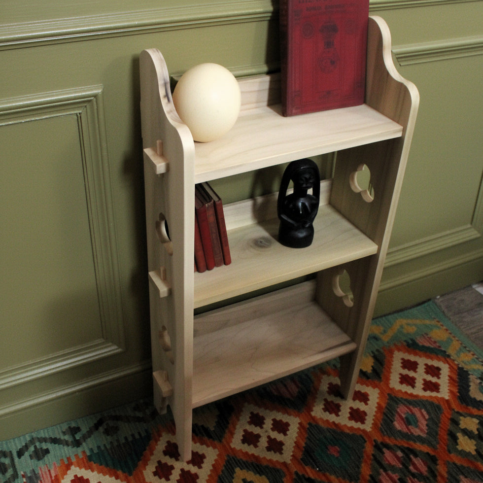 A small light wood bookshelf with three shelves, styled with a cream sphere, a red book, small red books, and a black decorative object, set against a cardboard box backdrop on a patterned rug.
