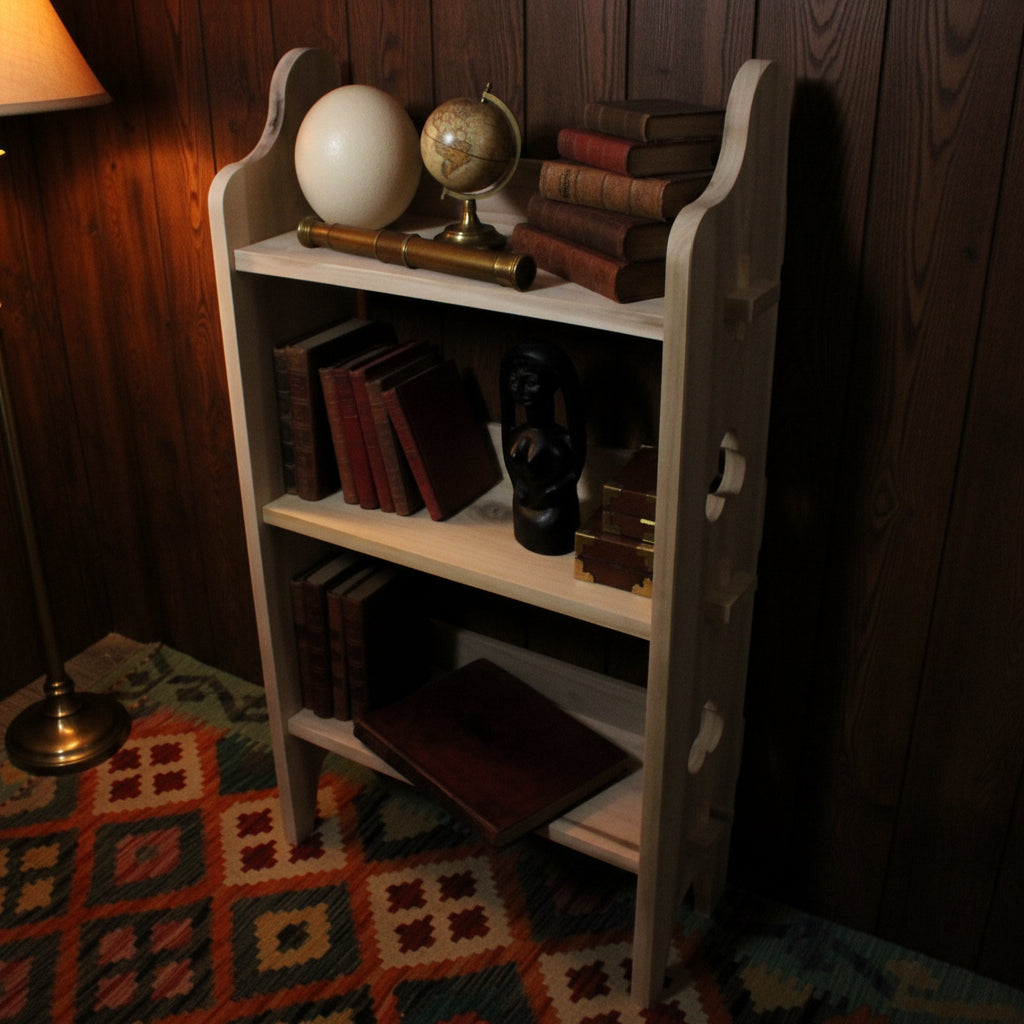 A small light natural wood three-tier bookshelf with decorative curved side supports, styled with books, a black mask, and a cream-colored sphere, set against a cardboard box background.