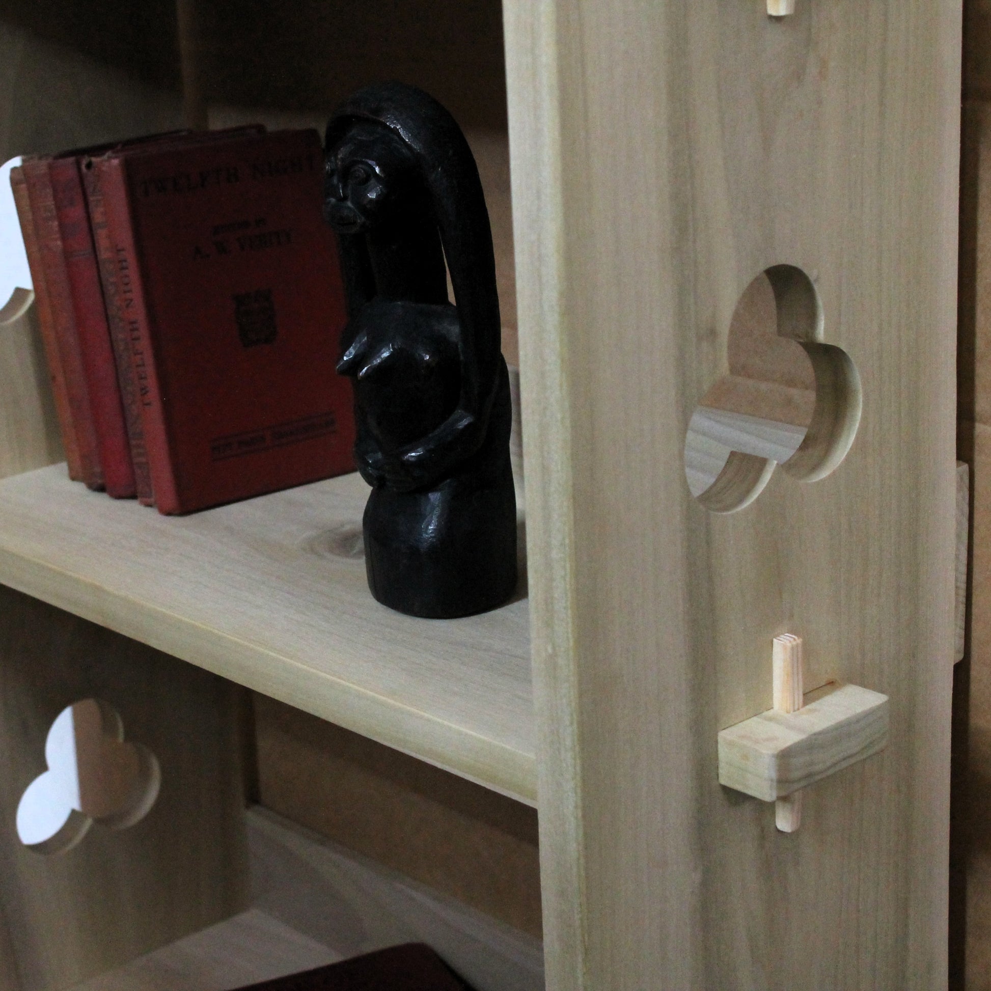Close-up of a light natural wood bookshelf or cabinet with decorative cutouts and a small wooden toggle detail, holding a black figurine and red books on the shelf.