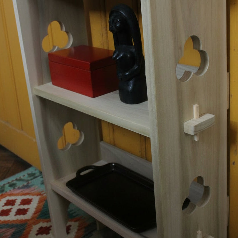 A light natural wood bookshelf with decorative cutout details on the side, holding a red book and a black figurine on the top shelf and a dark red book on the bottom shelf, set in an indoor room.