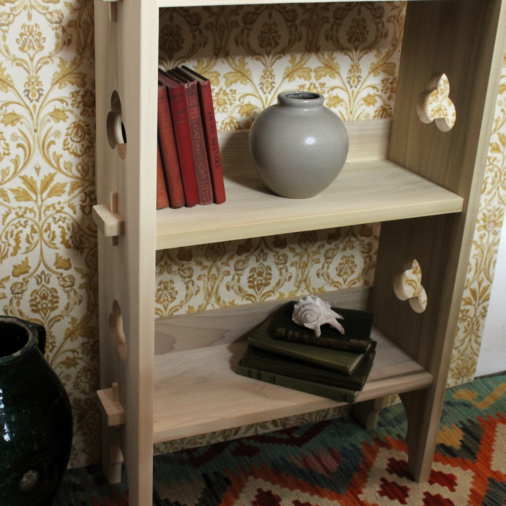 A light natural wood bookshelf with decorative cutout details on the sides, shown holding books and a glass vase in a home setting.