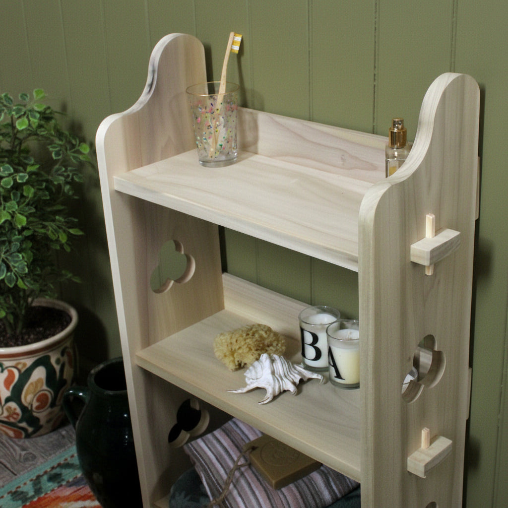 A light natural wood three-tier bathroom shelf with decorative cutouts, shown holding a toothbrush, small bottle, candle, sponge, shells, and folded towels.