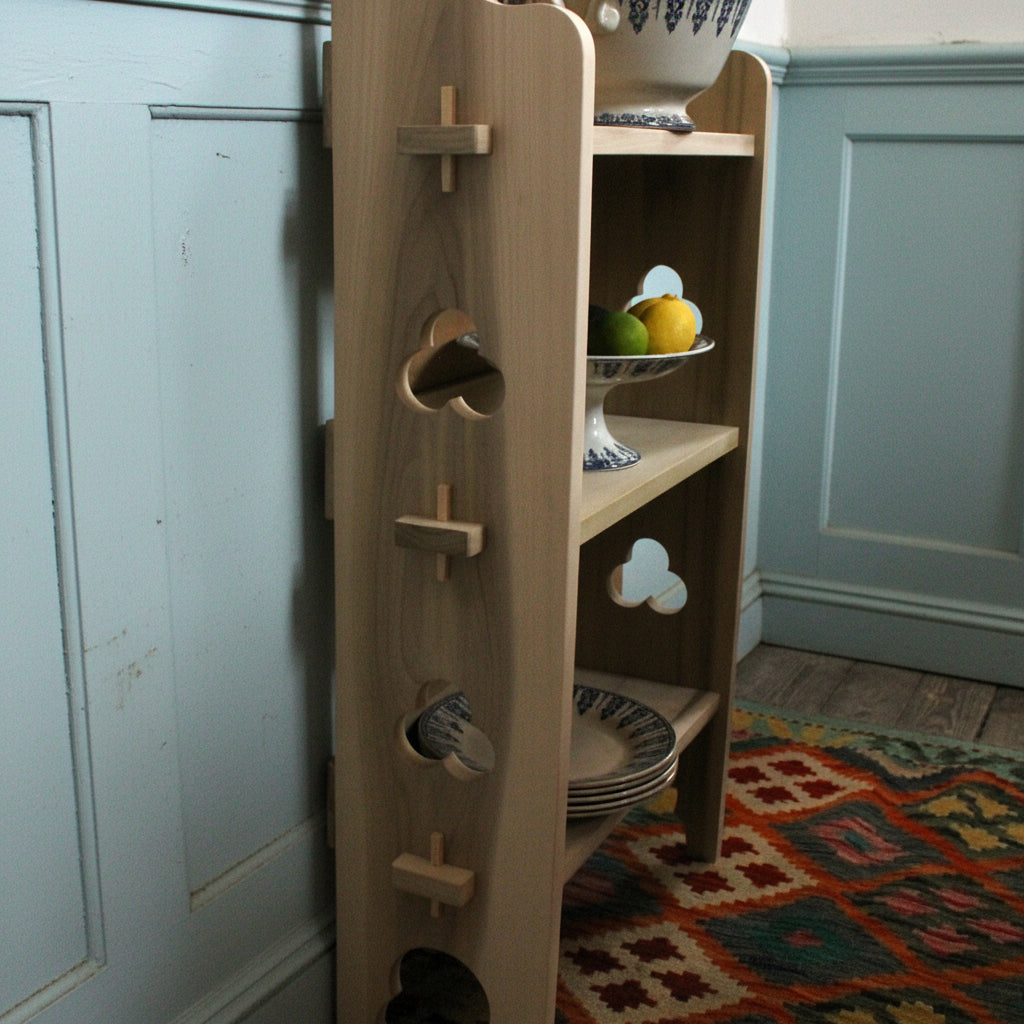 A light natural wood side table or shelving unit with three shelves, featuring decorative cutout shapes including hearts and clouds, shown in a workshop or room setting.
