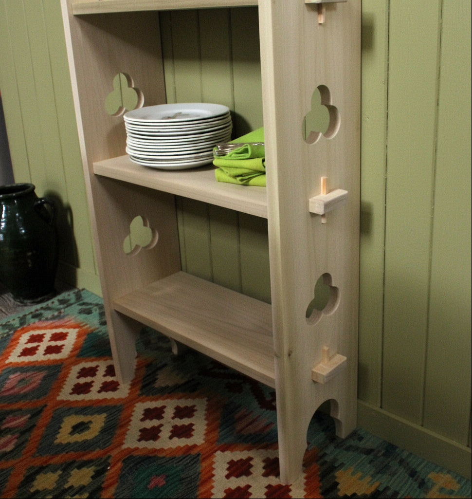 A light natural wood three-tier shelving unit with decorative cutout details on the sides, shown holding a stack of white plates and a green cloth, set on a patterned rug.