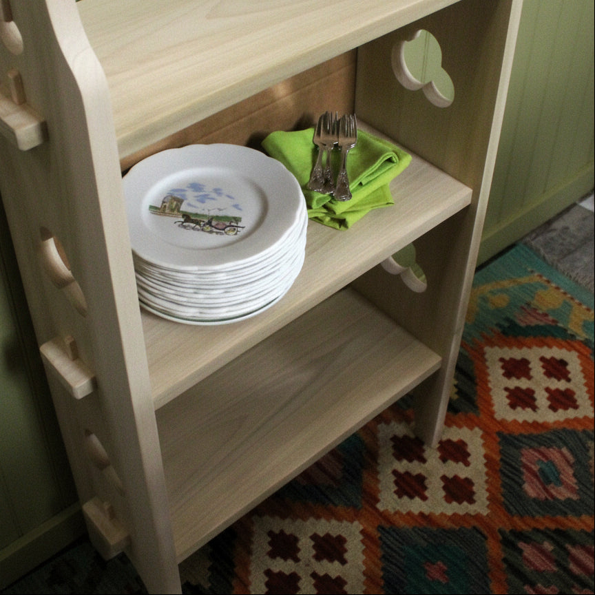 A light natural wood open shelving unit with three shelves, shown holding a stack of white plates, a green napkin, and cutlery. The shelf is positioned next to a cardboard box on a patterned geometric rug.