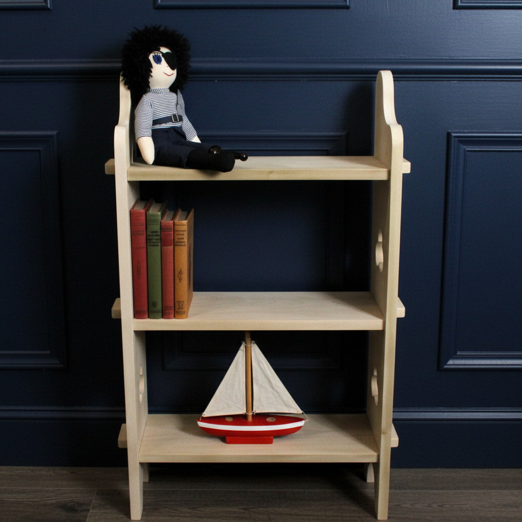 A light natural wood three-tier shelf with decorative curved side supports and small cutout details, shown standing indoors against a brown cardboard backdrop. A black-haired doll wearing a striped shirt and dark pants is placed on the top shelf.