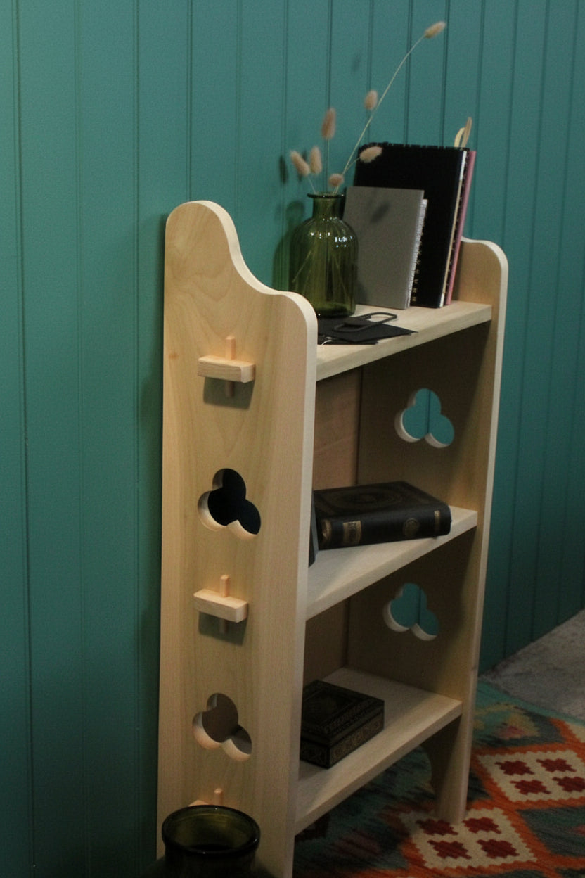 A light natural wood bookshelf with decorative cutout shelves, shown standing in a room with a large cardboard backing behind it, books on the shelves, and glass vases nearby.