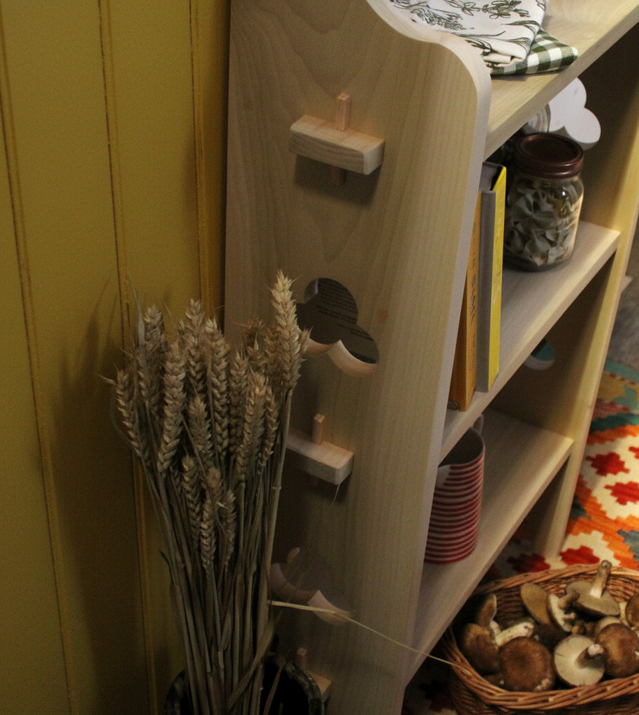 A light natural wood bookshelf with decorative cutouts and small wooden pegs, styled in a home setting next to dried wheat stems and a colorful patterned rug.