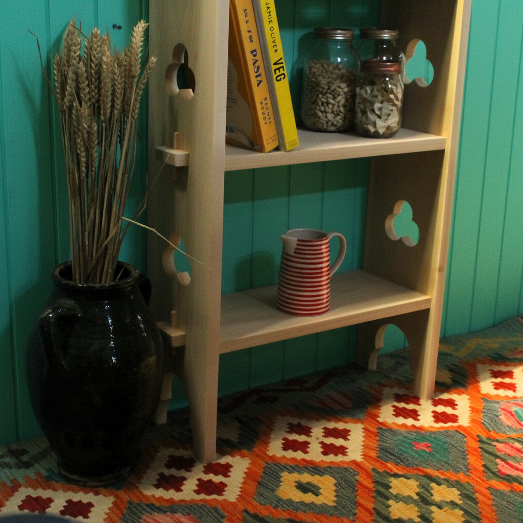A light natural wood three-tier shelf with decorative cutout details, styled with books, jars, and a red-and-white striped mug, next to a black vase with dried wheat on a patterned rug.