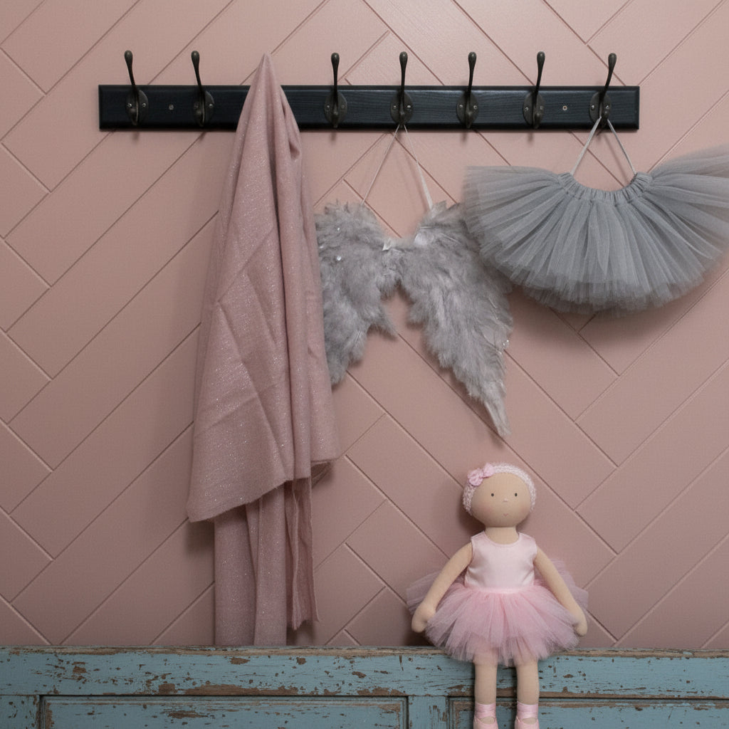 A black wall-mounted multi-hook coat rack with several dark metal hooks, shown against a plain off-white wall. A dusty pink towel hangs from one hook on the left, and a pair of gray feather angel wings hangs from a hook in the center-right.
