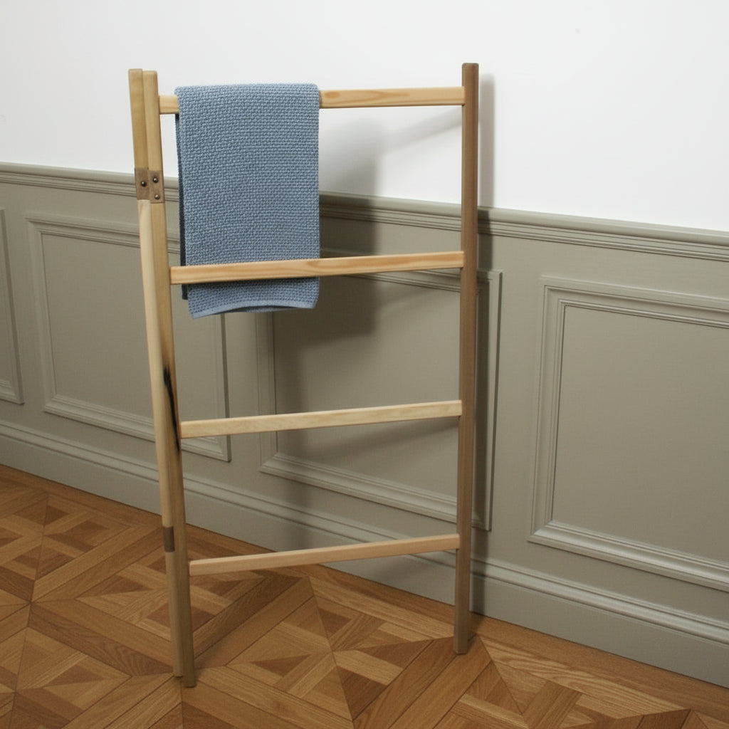 A light natural wood folding clothes rack with four horizontal bars, leaning against a white wall in a room with a wooden floor. A blue and white striped towel is draped over the side.
