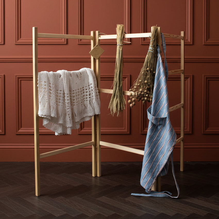 A light natural wood clothesline stand set outdoors in front of a dark green door, with a white knit sweater draped on the left, a bundle of dried grasses in the center, and a blue striped apron hanging on the right.