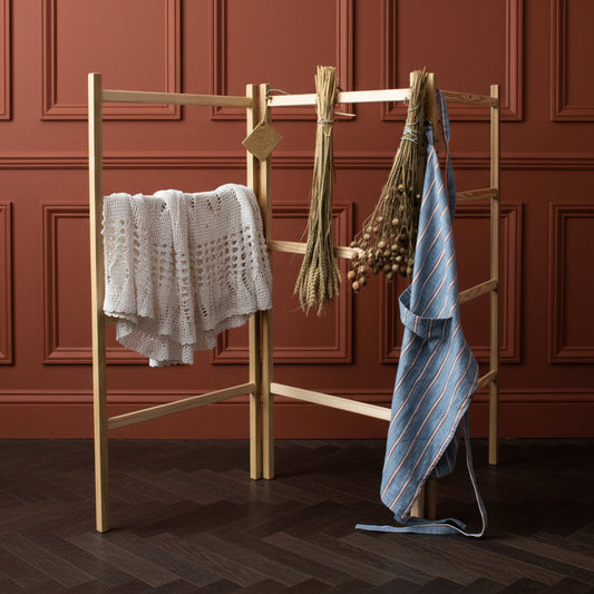 A light natural wood clothesline stand set outdoors in front of a dark green door, with a white knit sweater draped on the left, a bundle of dried grasses in the center, and a blue striped apron hanging on the right.