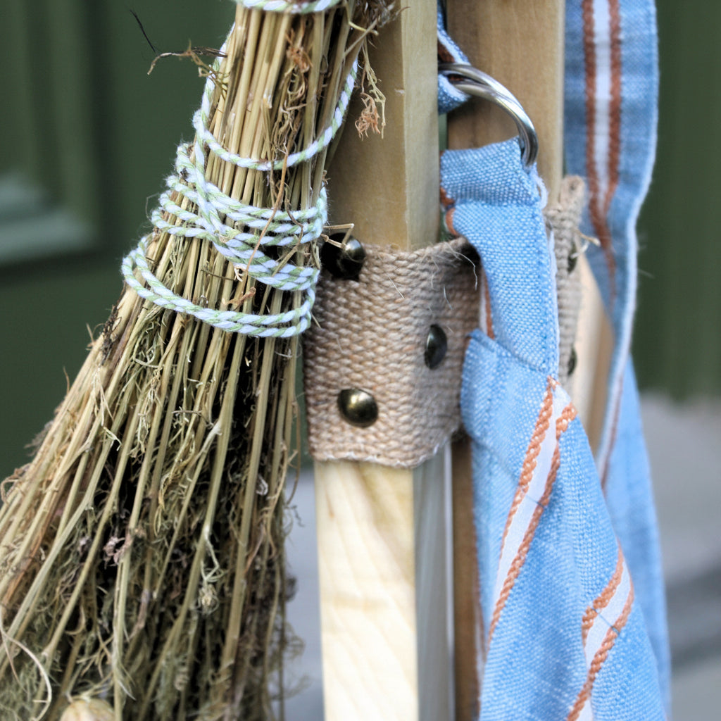 A close-up of a rustic decorative hanging arrangement featuring a small straw broom wrapped in white string, a light wood post, a beige burlap strap with metal snaps, and a blue fabric strip with orange stripes.