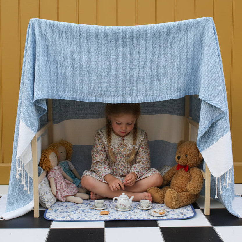 A litle girl having tea with dollies and a tedy, under a wooden clothes horse with a blue blanket thrown under it