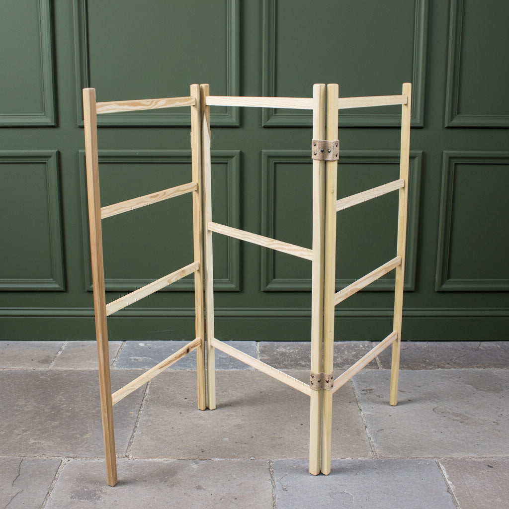 A light natural wood clothes drying horse with jute hinges, standing on a stne floor in front of a dark green wall panelling.